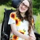 A smiling young woman with cerebral palsy, wearing glasses and a sunflower-print dress, sits in her wheelchair outside.